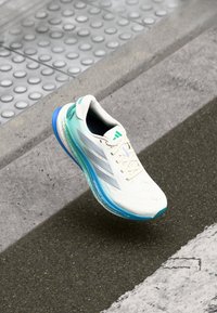 Baskets de course blanches avec des accents bleus et aqua, un supérieur en maille texturée, un design à trois bandes et une semelle rembourrée, positionnées au-dessus d'un trottoir.