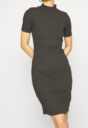 Kobieta w dopasowanej, prążkowanej, ciemnoszarej sukience o krótkich rękawach i długości do kolan, z półgolfem, stojąca z rękami za plecami.