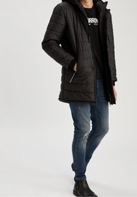 Černá péřová bunda s kapucí, prošívaný design, kapsy na zip a elastické manžety, nošená přes černou mikinu a modré džíny s efektem opotřebení.