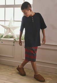 Marineblaues Kurzarm-Henley-Shirt mit dreiknopfiger Leiste, kombiniert mit rotkarierten Shorts und braunen Slippern. In einem Innenraum nahe einer Stern-Dekoration.