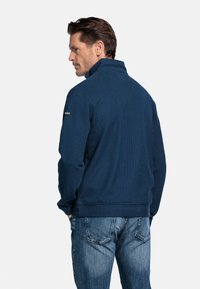 Marine blauwe pullover met ritskraag, gemaakt van een gestructureerde katoenmix, met ribboorden aan de manchetten en onderrand, gedetailleerd met een logopatch op de mouw. Jeans worden eronder gedragen.