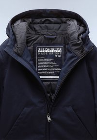 Veste matelassée noire avec capuche, partiellement zippée, montrant l'étiquette intérieure avec la marque Napapijri et un texte sur le matériau en polyester recyclé.