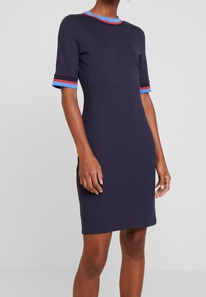 Mujer con un vestido azul marino hasta la rodilla, de mangas cortas y con ribete a rayas azules y rojas en el cuello y los puños.