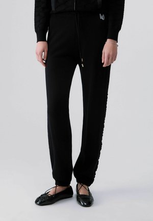 Personne portant un pantalon de jogging noir avec des poignets élastiques et des ballerines noires avec des lanières croisées, debout sur un fond clair.