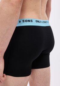 Άνδρας που φοράει μαύρα μπόξερ με ανοιχτό γαλάζια ζώνη μέσης που γράφει "ONLY & SONS", εμφανίζεται από τη μέση έως το μέσο του μηρού από πίσω.