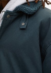 Närbild på en mörkgrön jacka med en stickad krage, tryckknapp och grå huvlinningsfoder synligt under.