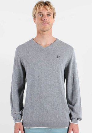 Homme blond portant un pull gris clair à col en V avec un petit logo noir sur la poitrine gauche, debout devant un fond blanc uni.