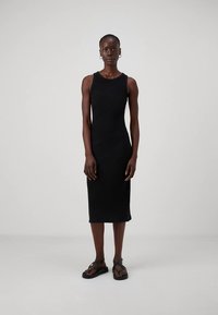 Moteris vilkinti juodą be rankovių midi suknelę ir juodas dirželines basutes, stovinti prieš šviesiai pilką fono.