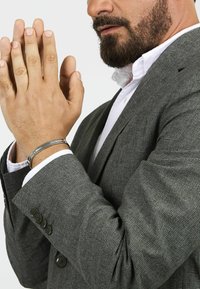 Bracciale in metallo argentato su una mano, abbinato a una giacca elegante grigia con texture e un sottile motivo a scacchi e bottoni neri.