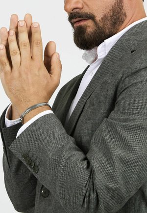 Bracciale in metallo argentato su una mano, abbinato a una giacca elegante grigia con texture e un sottile motivo a scacchi e bottoni neri.