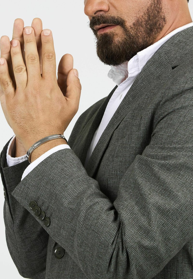 Bracciale in metallo argentato su una mano, abbinato a una giacca elegante grigia con texture e un sottile motivo a scacchi e bottoni neri.