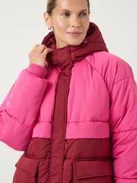Ženska, oblečena v puffer jakno v roza in burgundski barvi s kapuco, ki se dotika ovratnika in gleda na stran.