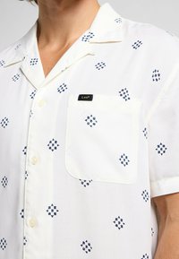 Kortärmad skjorta i lättviktig vit tyg med blå geometriska mönster, med en bröstficka och en varumärkesetikett.