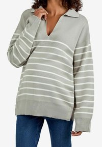 Femme portant un pull gris clair à col avec des rayures blanches horizontales et un jean bleu, tenant le col près de son cou avec une main.