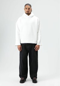 Sweat à capuche blanc oversize avec un col montant et des épaules tombantes, associé à un jean noir ample et des chaussures noires avec des lacets roses.