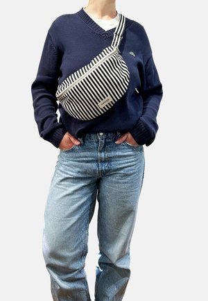 Personne portant un pull bleu marine, un jean bleu clair et un sac bandoulière rayé noir et blanc avec fermeture éclair et étiquette. Mains dans les poches.