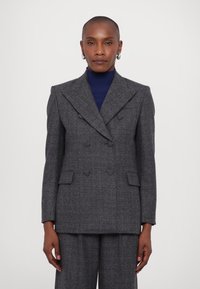 Grauer doppelt geknöpfter Blazer mit strukturiertem Schnitt, der einen strukturierten Stoff, spitze Revers und Fronttaschen aufweist. Kombiniert mit einem navyfarbenen Rollkragenpullover.