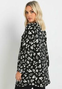 Tunic nera floreale con maniche lunghe, caratterizzata da motivi a fiori bianchi e una vestibilità comoda. Stoffa morbida con una leggera lucentezza.