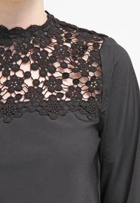 Blouse noire avec des détails en dentelle florale complexe au niveau de l'encolure et des épaules, avec un tissu lisse s'étendant le long des manches.