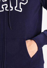 Sudadera navy con cremallera y letras blancas. Cuenta con capucha con cordón, bolsillos frontales y puños acanalados. Fabricada con una tela suave y texturada.