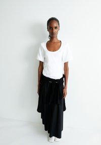 Weißes kurzärmliges Oberteil mit Rundhalsausschnitt, schwarzem Band in der Taille und langem schwarzen Rock. Das Modell trägt weiße Stiefel. Schlichtes Hintergrund.