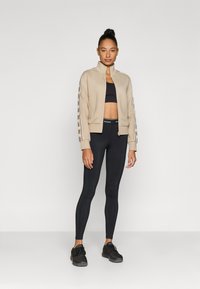 Μπεζ ζακέτα με φερμουάρ και μαύρες λεπτομέρειες με λογότυπο στα μανίκια, συνδυασμένη με μαύρο κοντό τοπ και μαύρα κολάν. Το μοντέλο φοράει μαύρα αθλητικά παπούτσια.