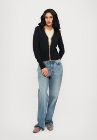 Cardigan navy con un design aderente, caratterizzato da una chiusura con zip e dettagli multicolore. Abbinato a jeans a gamba larga blu chiaro e scarpe a fantasia.