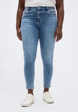 Άτομο που φοράει ανοιχτό μπλε στενά τζιν και λευκά αθλητικά παπούτσια, στέκεται μπροστά σε απλό φόντο.