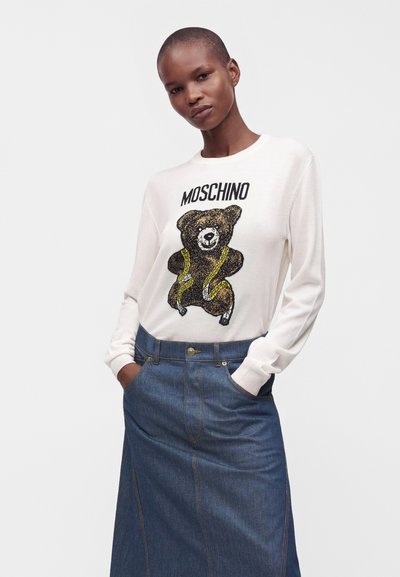 Femme portant une chemise blanche à manches longues avec un ours en peluche et le texte "MOSCHINO", associée à une jupe en jean bleue taille haute.