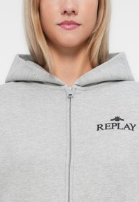 Sivi zip-up dukserica napravljena od mješavine pamuka, s prednjim patent zatvaračem i crnim "REPLAY" logotipom iznad grafike krune. Teksturalna tkanina.