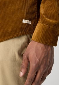 Camisa de pana marrón con textura acanalada y etiqueta de marca. La manga derecha presenta un puño grueso. Tela de color claro debajo.