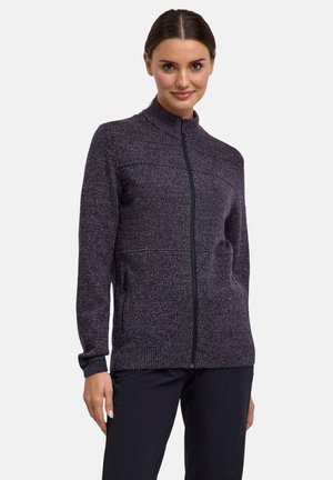 Femme portant un pull en maille zippé violet foncé avec col montant et manches longues, associé à un pantalon noir, debout devant un fond clair.