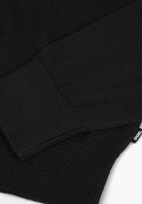 Detail eines schwarzen Strickpullovers mit gerippten Bündchen und Saum, glatter Textur und einem gewebten Markenetikett in der unteren Ecke.