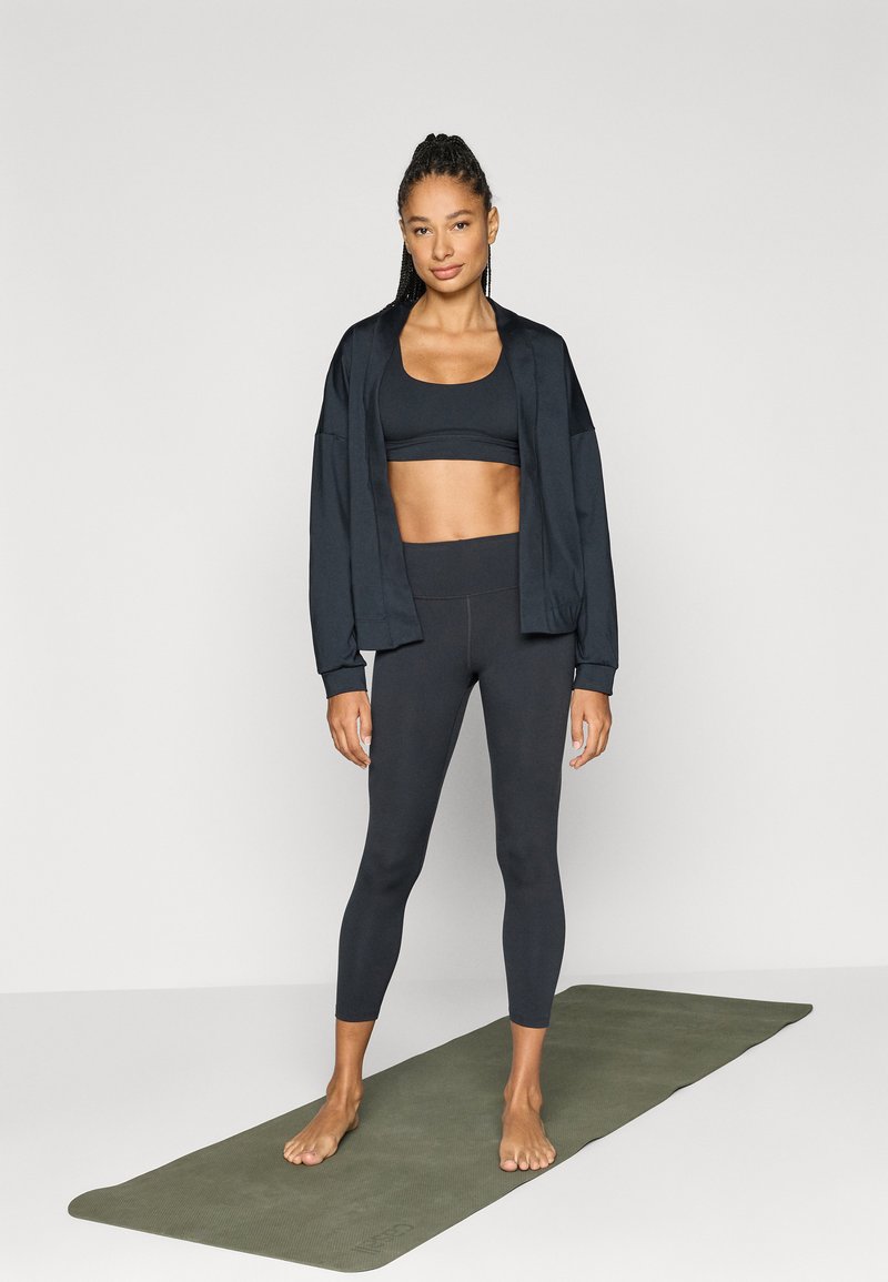 Zwart sportoutfit bestaande uit een nauwsluitende sportbeha, high-waisted leggings en een jack met een losse pasvorm, op een groene yogamat.