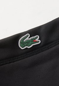 Um tecido preto com uma textura suave apresenta um logo de crocodilo verde contornado com fio cinza, posicionado perto de uma costura de um lado.