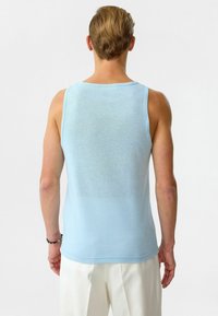 Lichtblauw, mouwloze tanktop met een subtiele textuur. Heeft een ronde halslijn en een ontspannen pasvorm aan de achterkant, gedragen met witte broeken.