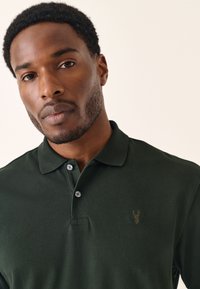Ciemnozielona koszulka polo z teksturowanej tkaniny, z dwoma guzikami na plisach i małym, wytłoczonym emblematem na lewej piersi.