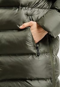 Kurtka puchowa w ciemnooliwkowym kolorze, z kieszenią na zamek błyskawiczny i pikowaną teksturą. Materiał jest błyszczący i gładki, o miękkim wyglądzie.