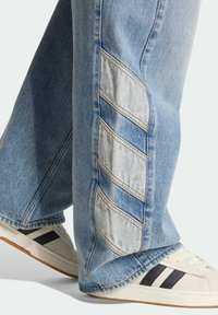 Ljusblå jeans i denim med kontrasterande paneler, med ett chevronmönster på sidorna. Bärs med vita sneakers prydda med svarta ränder.