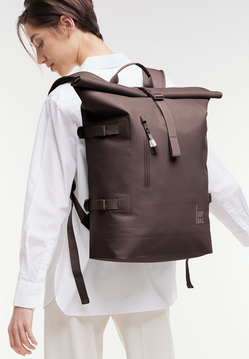 Mochila enrollable impermeable marrón con correas ajustables, cremallera en el bolsillo frontal y detalle del logo en la parte inferior derecha.