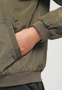 Olijfgroene bomberjack met een gladde, glanzende buitenkant, geribbelde manchetten en een zijzak. Voorzien van een elastische tailleband voor een aansluitende pasvorm.
