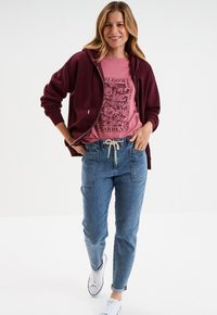 Felpa a zip maroon, t-shirt rosa con grafica a fiori, joggers in denim blu con vita elasticizzata e sneakers bianche.
