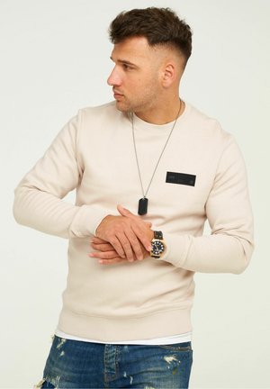Light beige sweatshirt with round neckline, ribbed cuffs, and a textured black logo patch on the chest. Worn with distressed blue jeans.