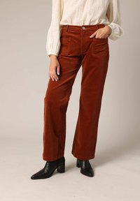 Pantalons à pattes larges en velours côtelé couleur rouille avec poches latérales, associés à un blouson crème clair et des bottines noires à talon épais.