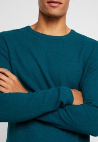 Man med korslagda armar som bär en strukturerad turkos tröja, visad från axlar till läppar mot en enkel ljus bakgrund.