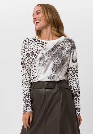 Smiling woman wearing a white leopard-print long-sleeve top and high-waisted dark brown leather skirt with a belt.