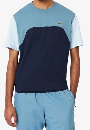 Homme portant un t-shirt à manches courtes bicolore bleu marine et bleu clair avec un short bleu clair assorti, mains relâchées le long du corps.
