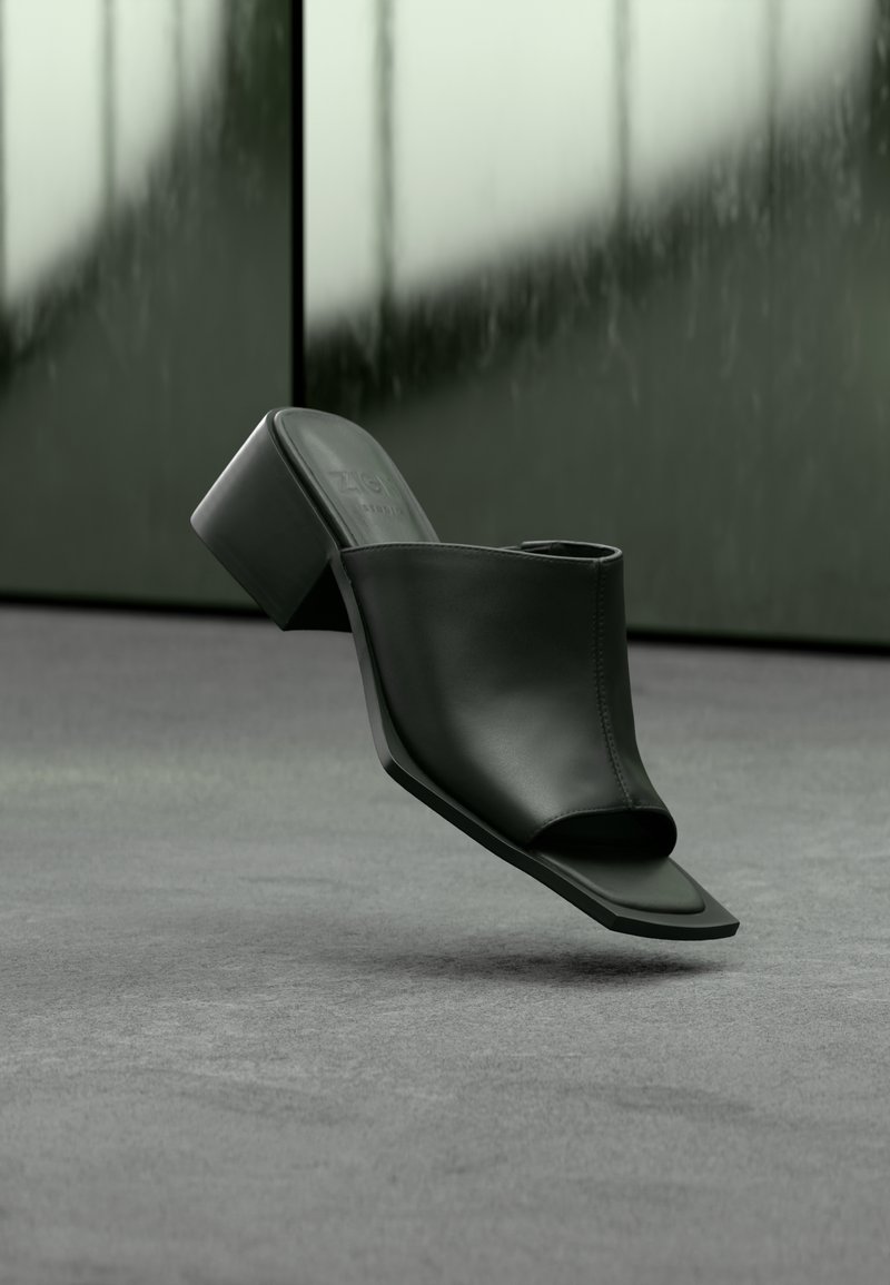 Mule noire en cuir à bout ouvert avec talon bloc, flottant au-dessus d'un sol moquetté gris avec un fond en verre flou.