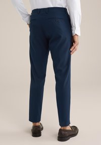 Homme vêtu de pantalons ajustés bleu marine, d'une chemise blanche et de mocassins en cuir marron foncé, vu de dos, se tenant sur un sol beige.