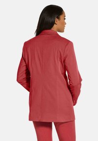 Roter maßgeschneiderter Blazer mit strukturierten Schultern, glatter Textur und Rückenschlitz. Mit langen Ärmeln und einer einzelnen Rückennaht.